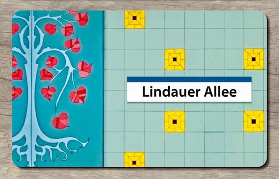 U-Bahnhof Lindauer Allee Frühstücksbrettchen - schwarze Schrift auf einem weißen Schild mit dunkelblauem Rand an der Oberkante, im Hintergrund mint-farbene Fliesen mit vereinzelt gelben Fließen, zu linken Seite eine abstrakte Linde in blau Tönen mit rot-r