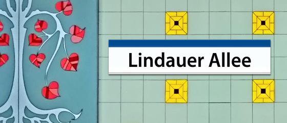 U-Bahnhof Lindauer Allee Frühstücksbrettchen - schwarze Schrift auf einem weißen Schild mit dunkelblauem Rand an der Oberkante, im Hintergrund mint-farbene Fliesen mit vereinzelt gelben Fließen, zu linken Seite eine abstrakte Linde in blau Tönen mit rot-r