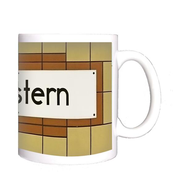 U-Bahnhof Südstern Kaffeetasse - weißes Schild mit schwarzer, schlichter Schrift auf weißem Schild mit rötlich-brauner Umrandung auf orange-gelben Fliesen