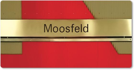 U-Bahnhof Moosfeld Kühlschrankmagnet - schwarze Schrift auf goldenem Schild, vor rotem Untergrund mit grünlichen Elementen