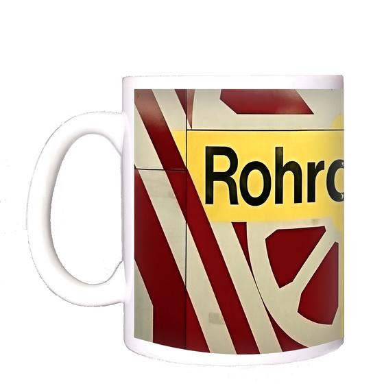 U-Bahn-Kaffeetasse Berlin Rohrdamm - schwarze Schrift auf gelbem Schild, umrandet von weißem, radähnlichem Muster auf rot-braunem Untergrund
