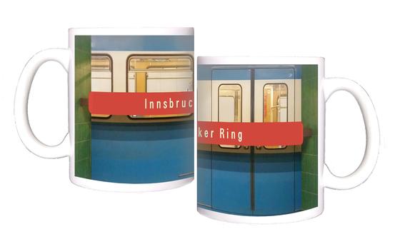 U-Bahnhof Innsbrucker Ring Kaffeetasse - weiße Schrift auf rotem Streifen, vor blauem U-Bahnwagon, zwischen grünen Säulen