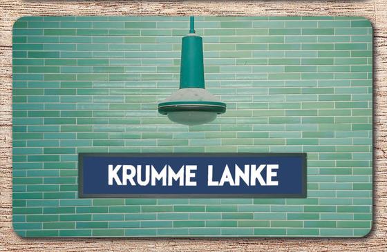 U-Bahnhof Krumme Lanke Frühstücksbrettchen - weiße Schrift auf blauem Schild, vor grün-blauer Fliesenwand, unter türkis-blauer Lampe