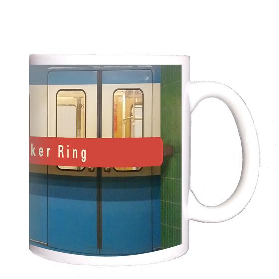 U-Bahnhof Innsbrucker Ring Kaffeetasse - weiße Schrift auf rotem Streifen, vor blauem U-Bahnwagon, zwischen grünen Säulen